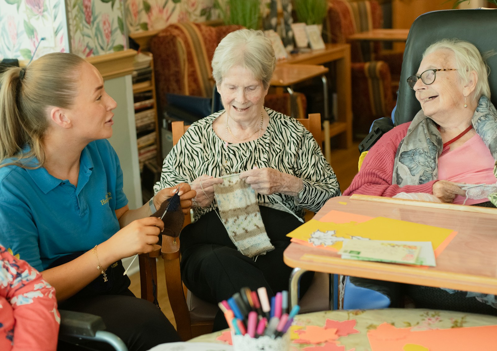 Betreuungskraft sitzt im Gemeinschaftsraum einer Pflegeeinrichtung bei zwei älteren Frauen und strickt mit ihnen